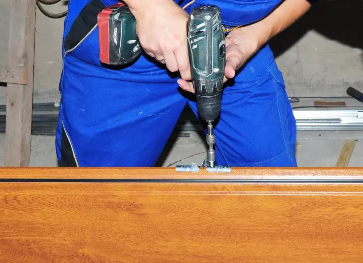 Worker uses cordless drill to install a hinge on a wooden cabinet in a workshop setting during daylight hours Garage Door repair Technician