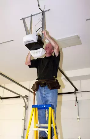 Man installs overhead garage door opener while standing on ladder in a garage during daylight hours